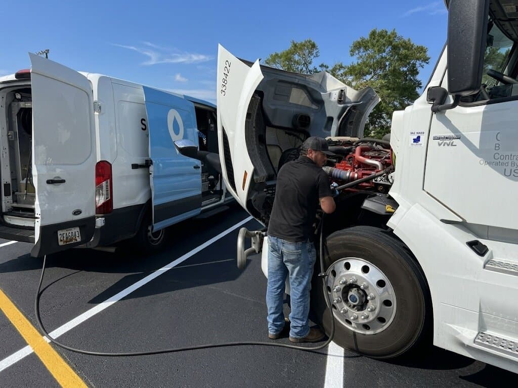 Slick technician servicing a Class 8 tractor on a customer yard beside a Slick service van under a clear blue sky